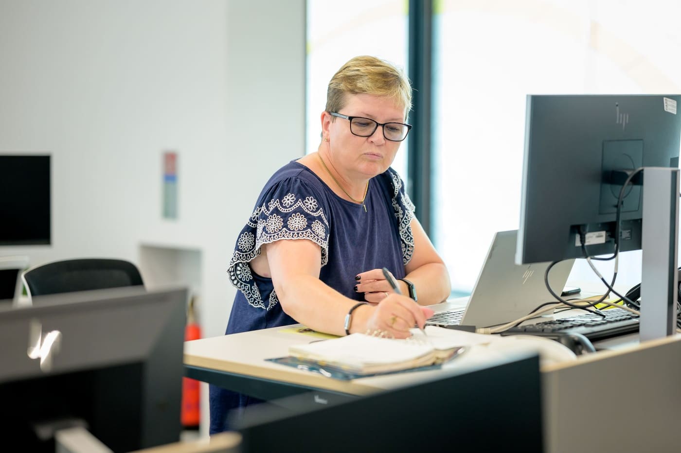 A woman in glasses takes notes at a standing desk beside a laptop and monitor