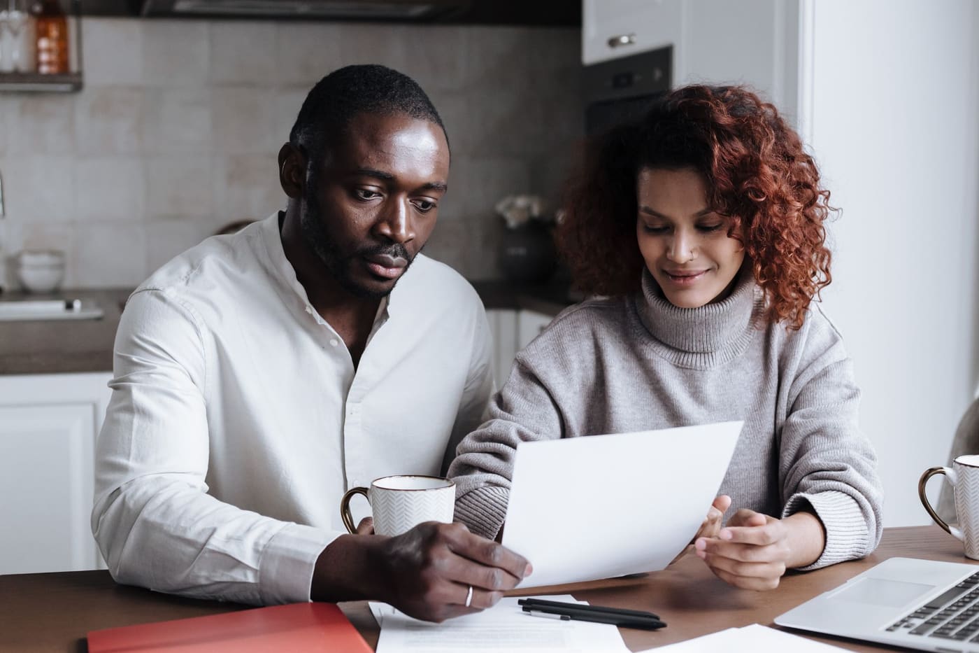 Two adults at a kitchen table review a printed document next to a laptop and coffee mugs