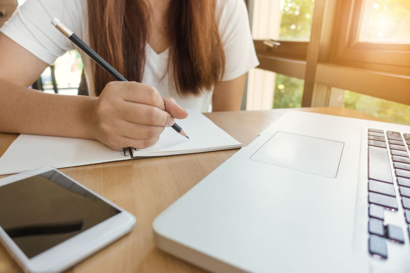 A person writing in a notebook with a pencil beside a laptop and a phone on a wooden desk
