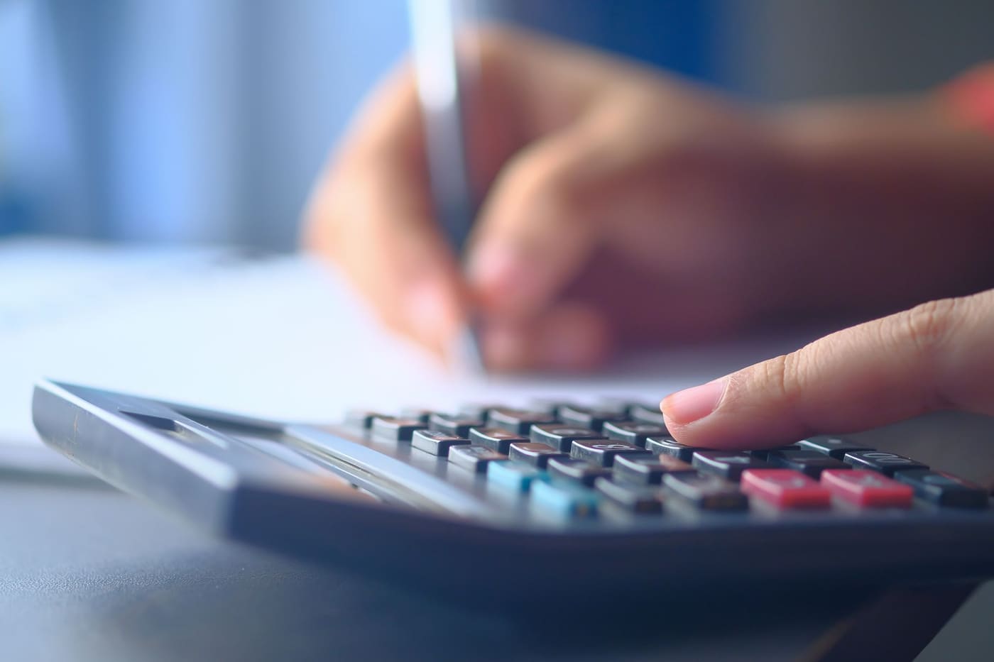 Close-up of a finger pressing calculator keys while another hand writes figures on paper