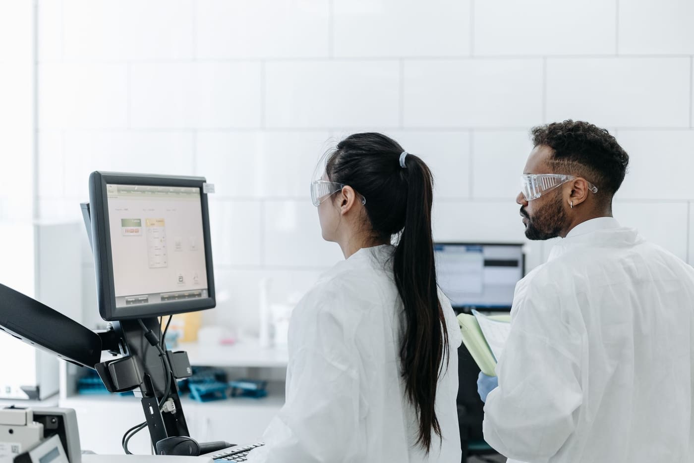 Two lab researchers in safety glasses and white coats reviewing results on a computer screen
