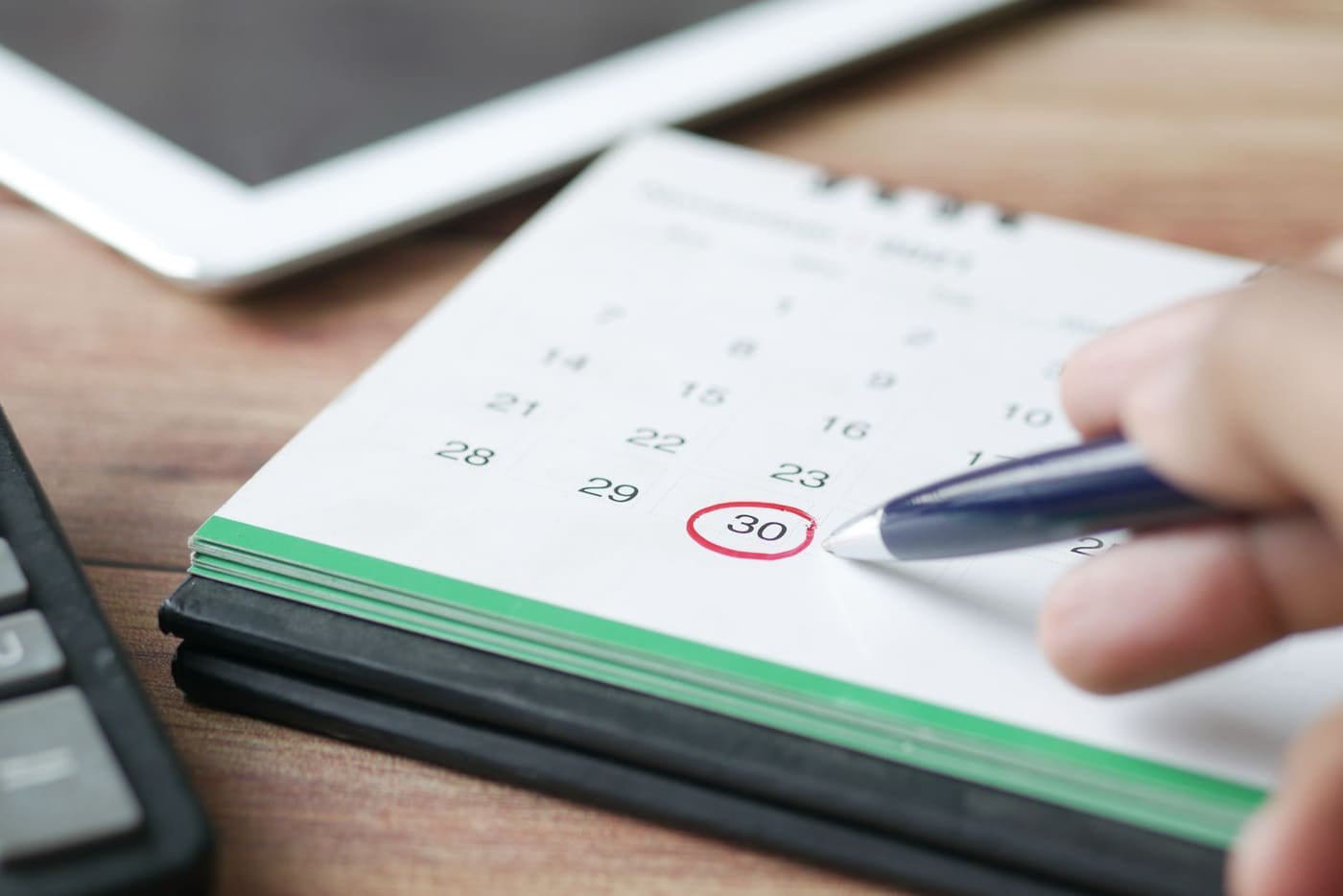 A hand holding a pen circles the 30th on a desk calendar beside a tablet
