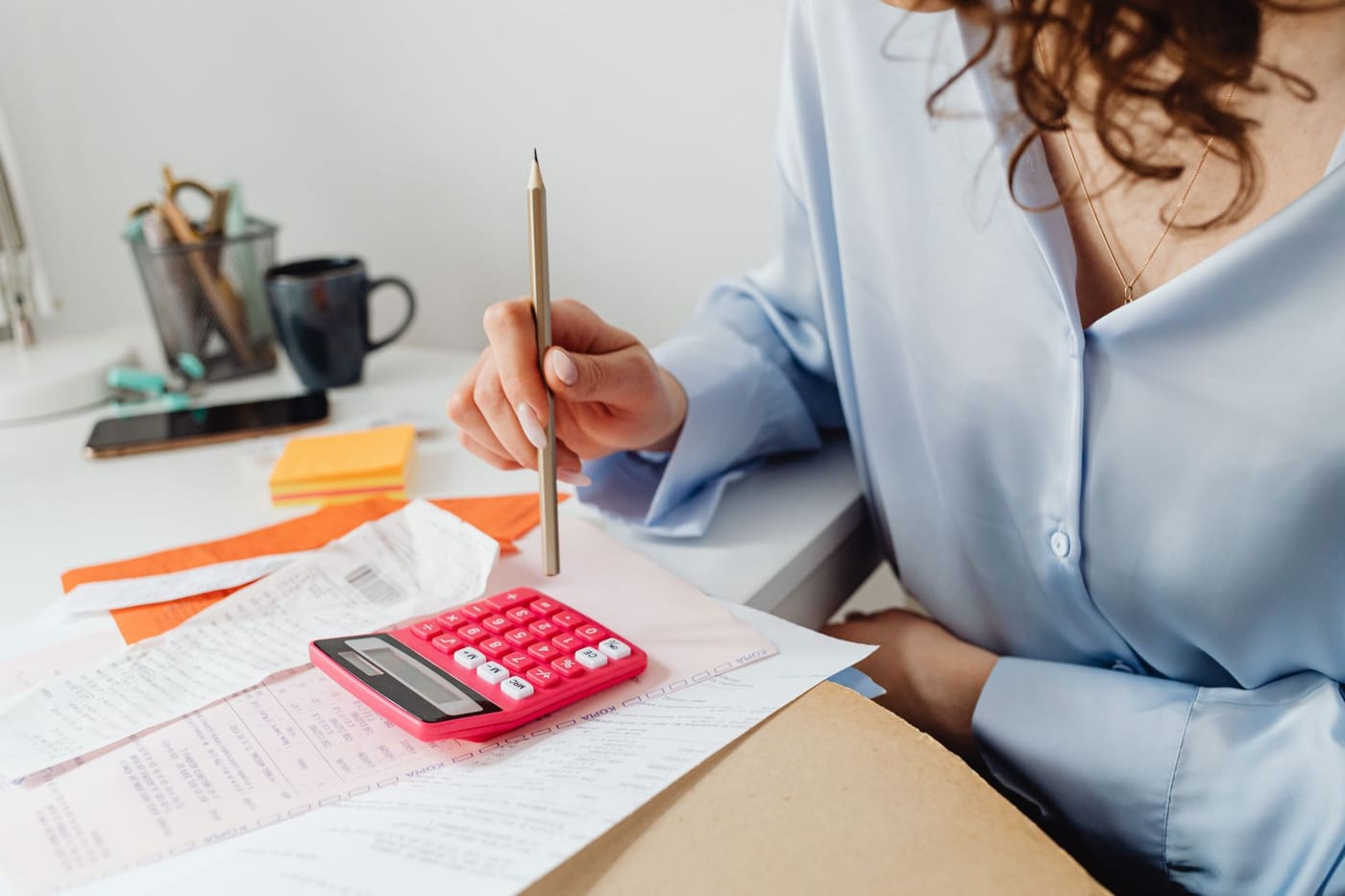 A woman holds a pencil over a pink calculator and a spread of bills and receipts on her desk