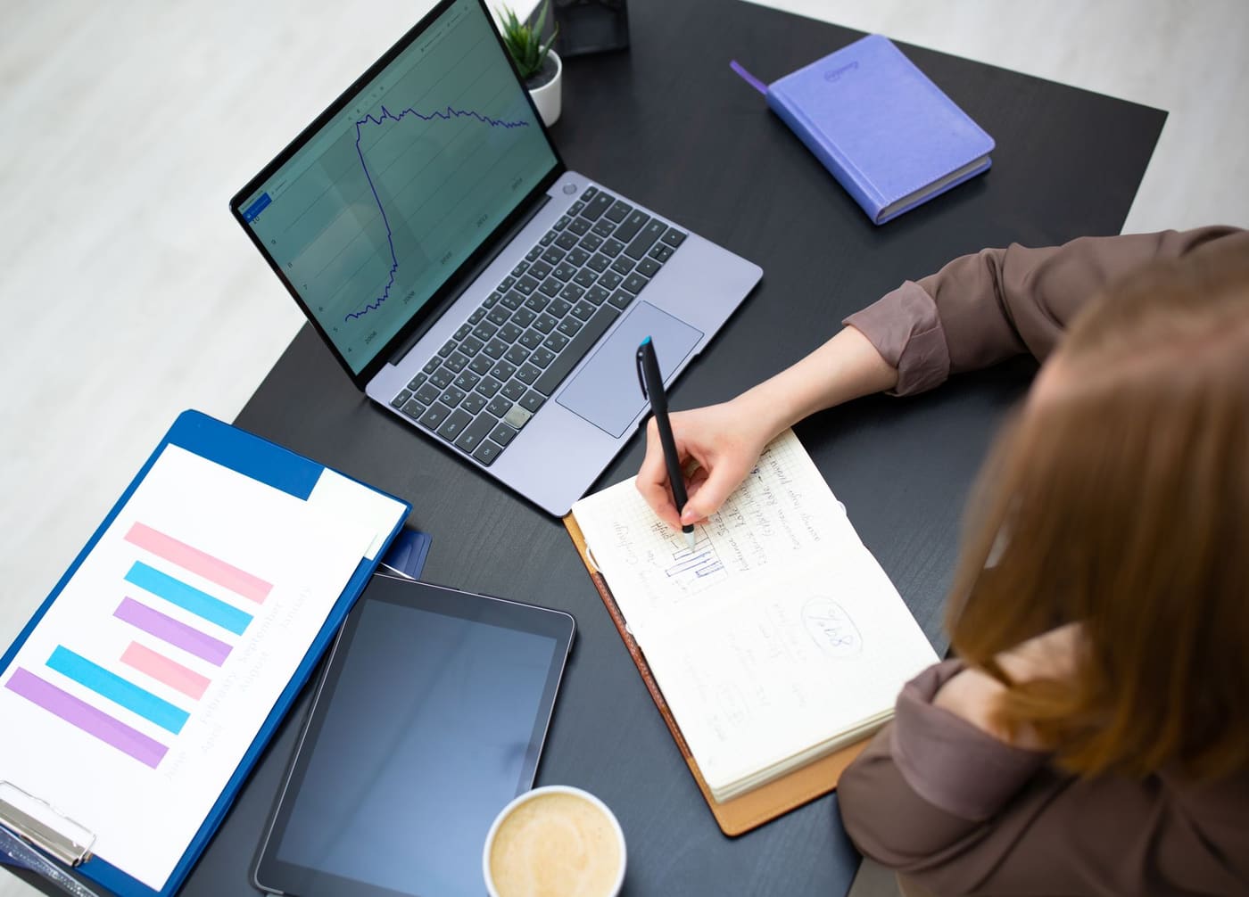 Overhead view of a woman writing notes beside a laptop showing a line graph and a printed bar chart on a clipboard