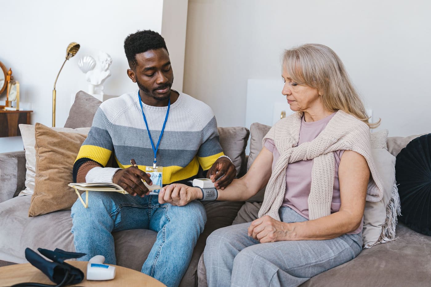 A community health worker with an ID lanyard checks an older woman's blood pressure with a wrist monitor on her living room sofa