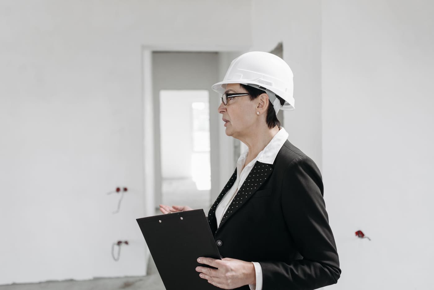 An inspector in a white hard hat and dark blazer holds a clipboard while walking through an unfinished building interior