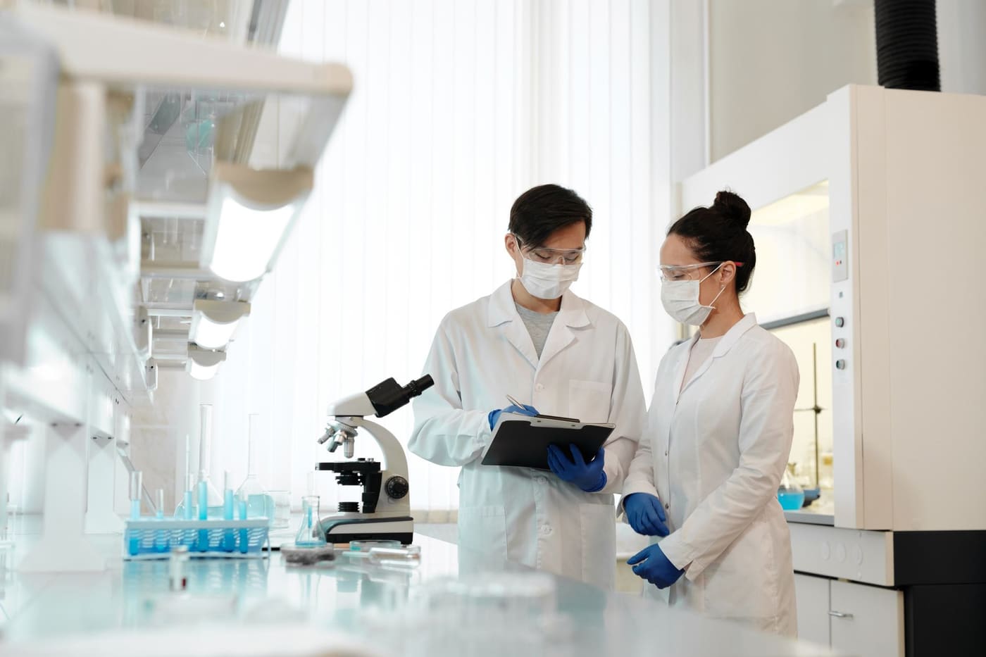 Two researchers in lab coats, masks and safety glasses review notes on a clipboard beside a microscope