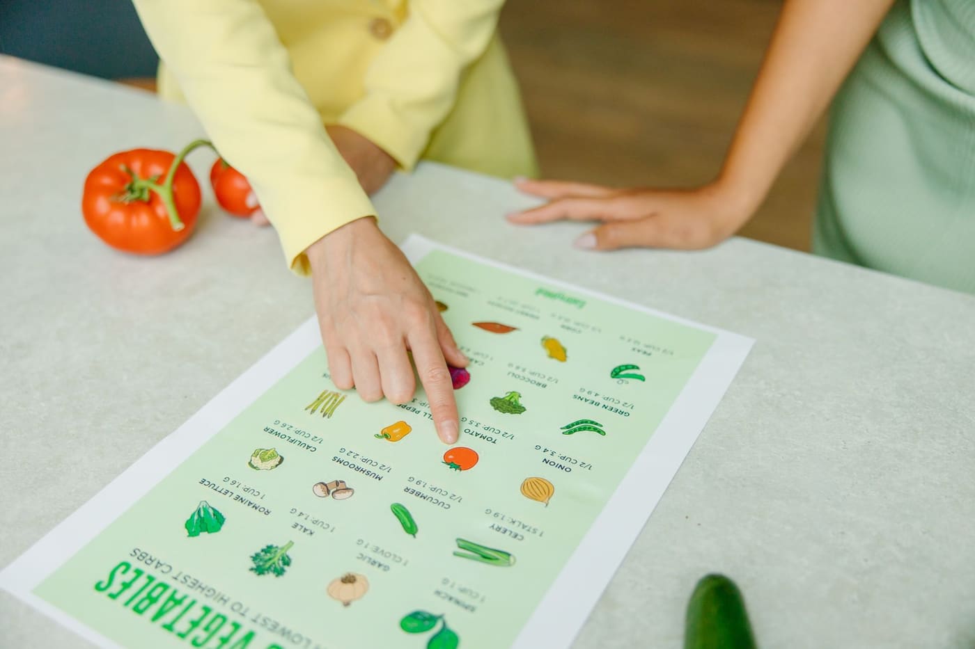 A hand points to a tomato on an illustrated vegetable nutrition chart while another person listens across the table