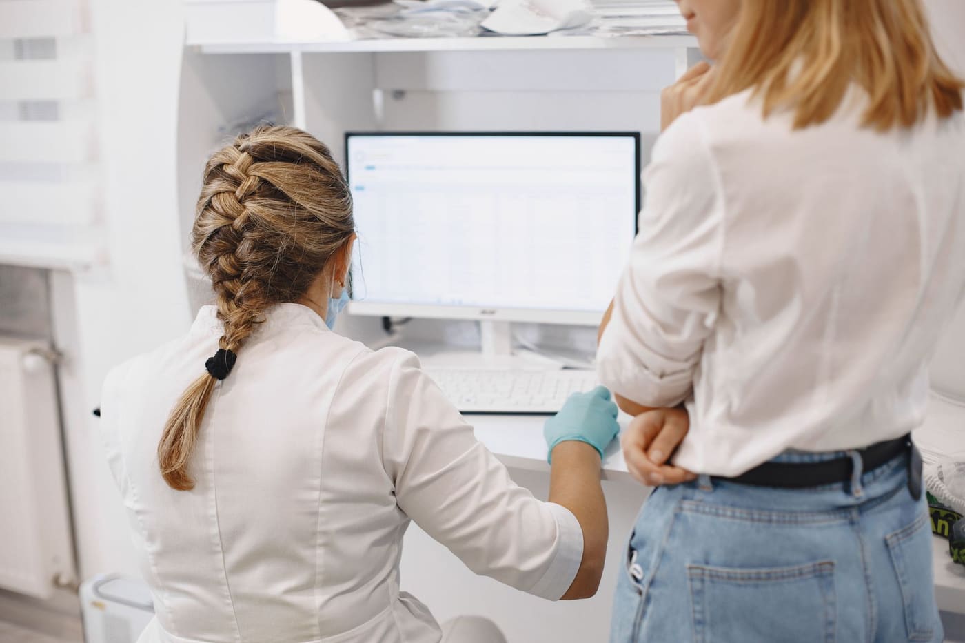 A clinician in a white coat and gloves works at a computer showing a data table while a colleague looks on