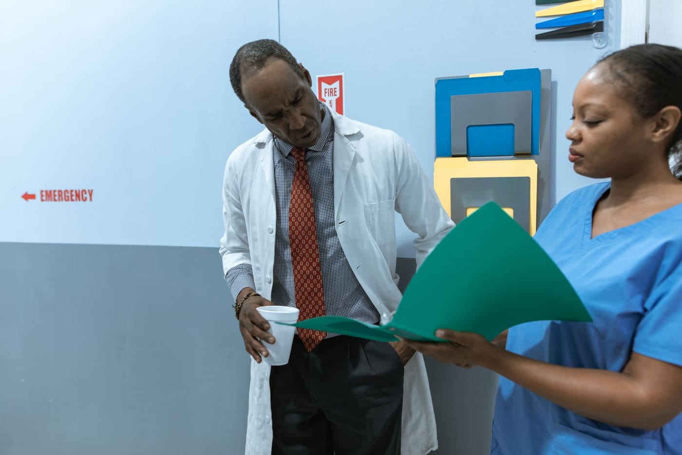 A physician in a white coat and a nurse in blue scrubs review a green folder together in a hospital corridor