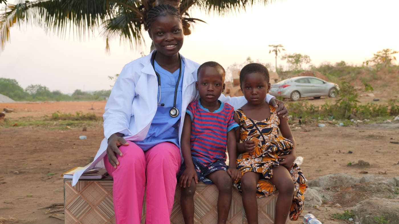 A health worker in a white coat and stethoscope sits outdoors with an arm around two young children