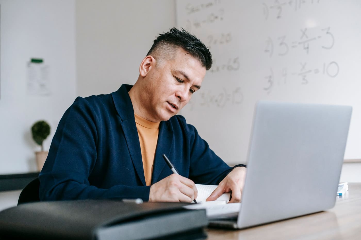 A man in a navy jacket writes in a notebook beside an open laptop, with math equations on a whiteboard behind him