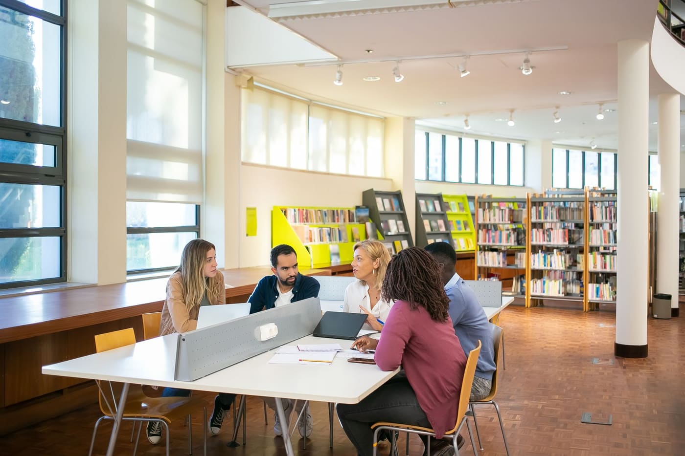 Five graduate students talk around a shared table with laptops and notebooks in a bright library