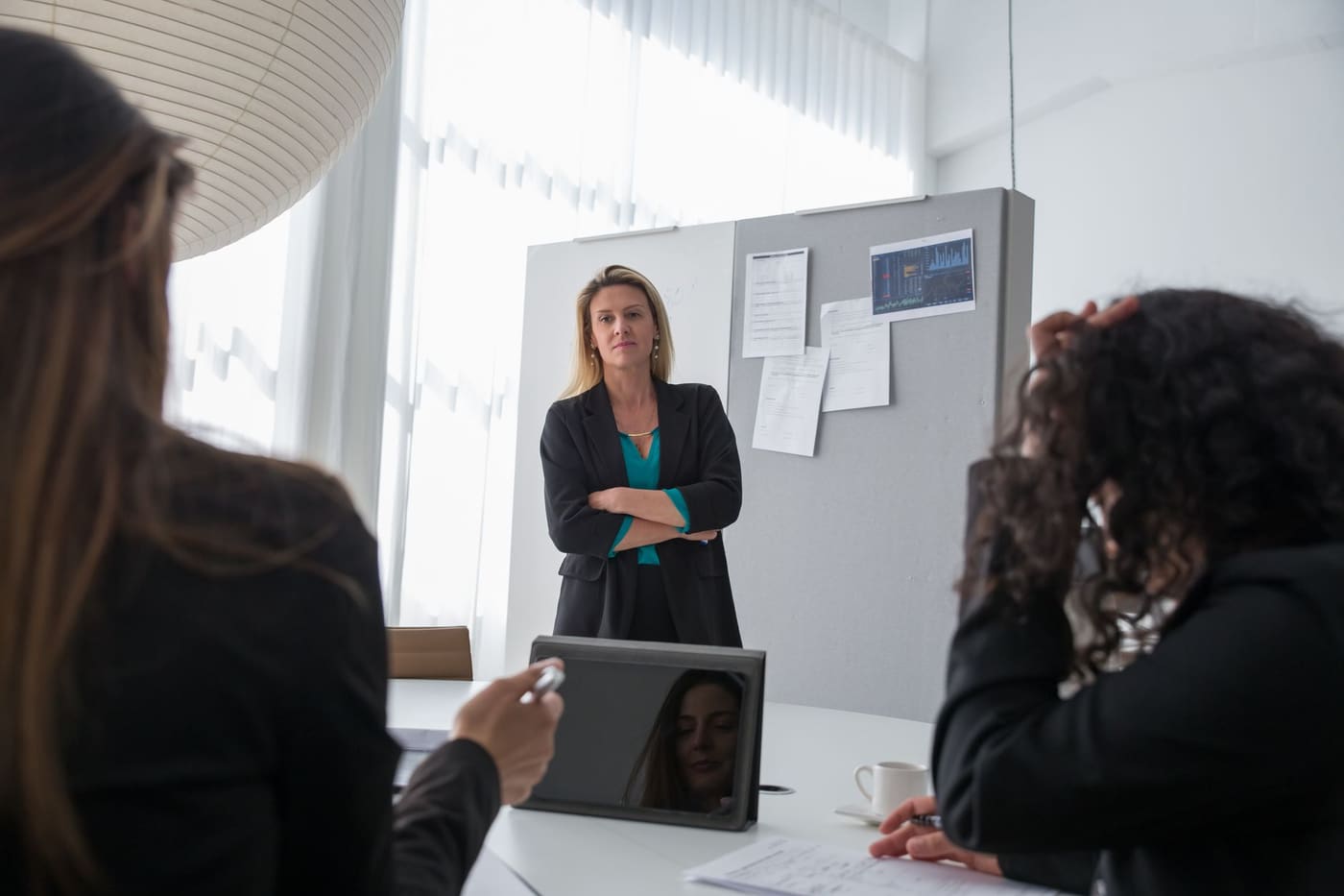 A woman in a dark blazer stands with arms folded at the head of a conference table, leading a meeting in front of a board of charts