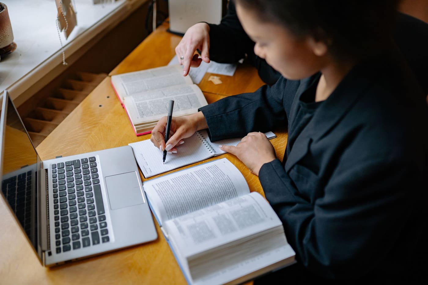 Two students at a wooden desk with open textbooks, a notebook and a laptop, one writing notes while the other points at a page