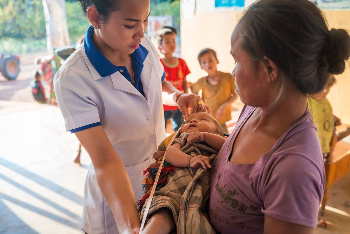 A health worker in a white uniform measures an infant held by its mother at a rural clinic in Laos while children watch