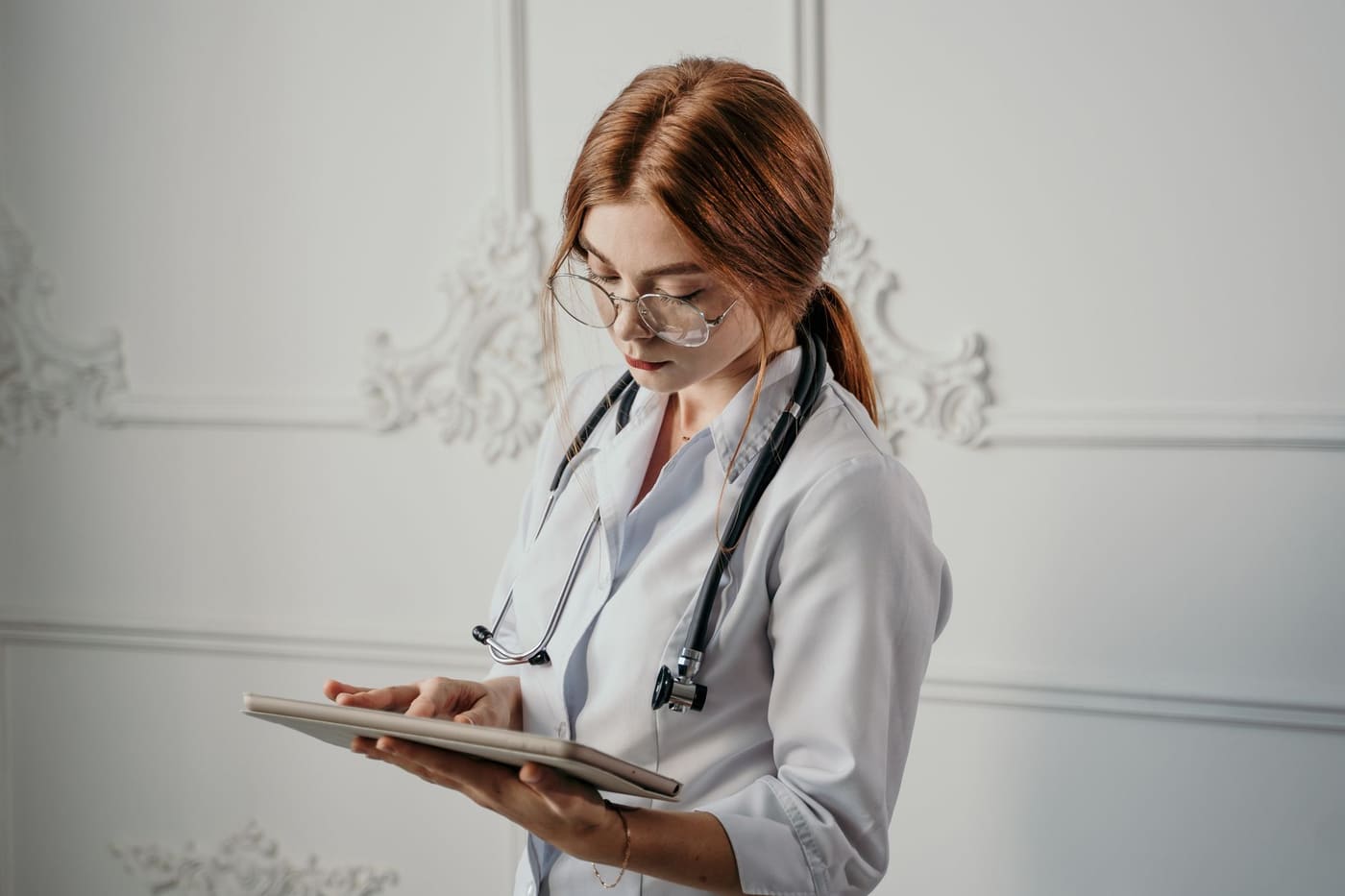 A physician in glasses and a white coat with a stethoscope reviews patient information on a tablet
