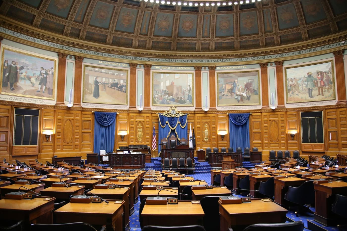 Empty chamber of the Massachusetts House of Representatives with rows of wooden desks, microphones and murals below a painted dome
