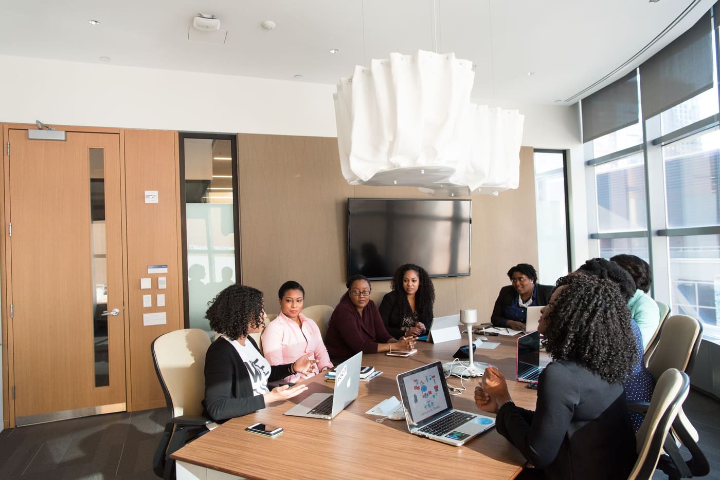 Eight women with laptops talk around a long conference table in a bright meeting room