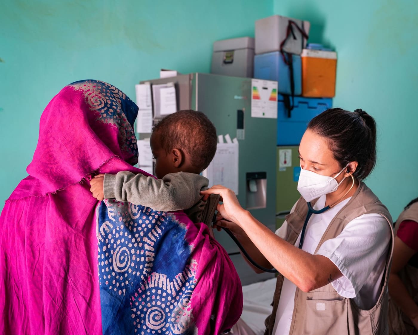 A health worker in a mask listens with a stethoscope to a toddler held by a mother in a bright pink wrap at a clinic