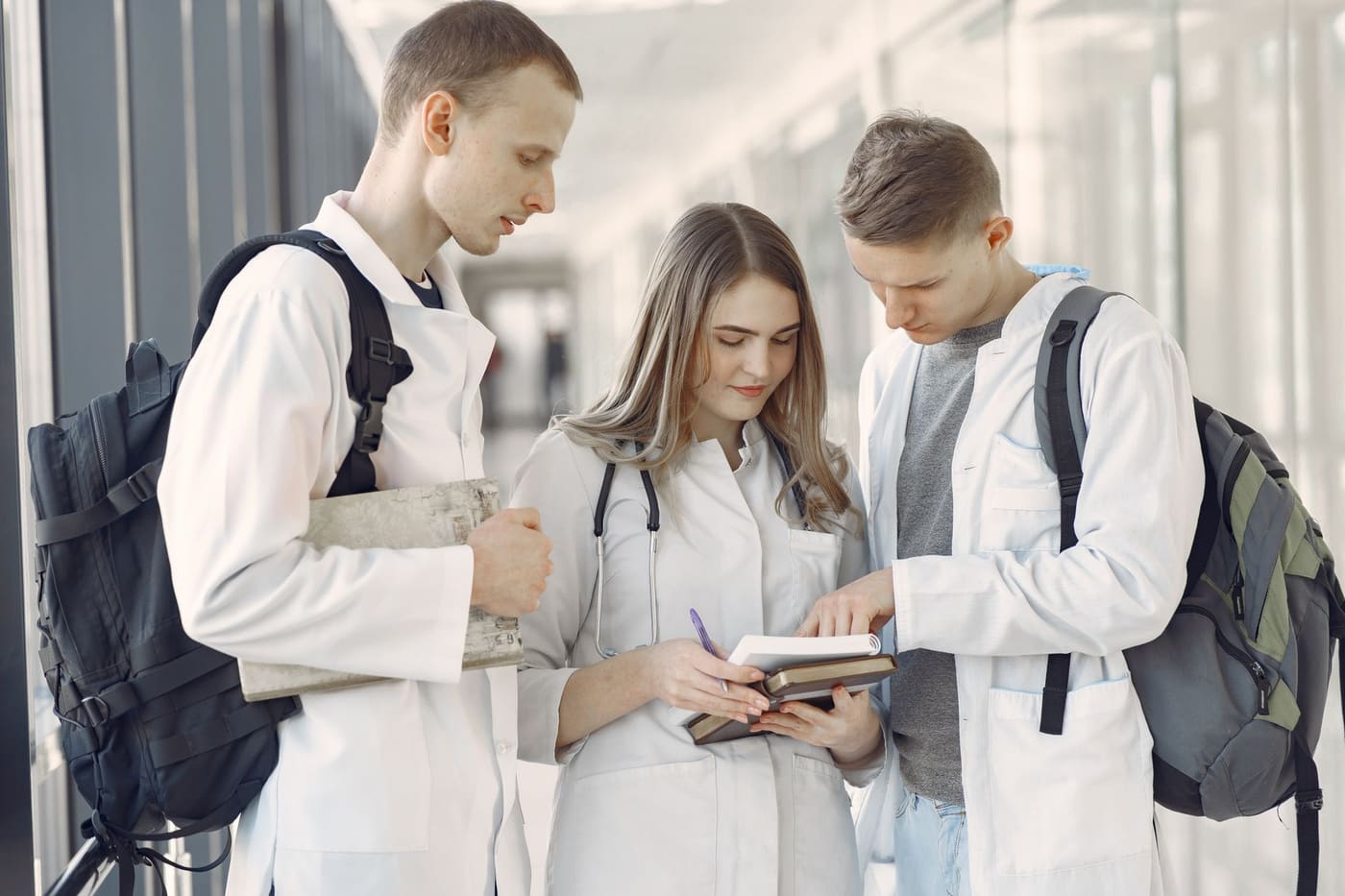 Three medical students in white coats with backpacks review a notebook together in a bright hallway
