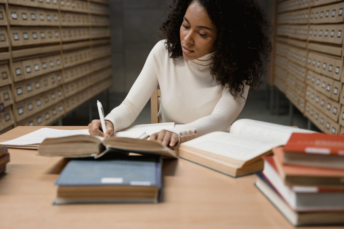 A woman in a cream sweater takes notes from open books at a library table lined with archive drawers