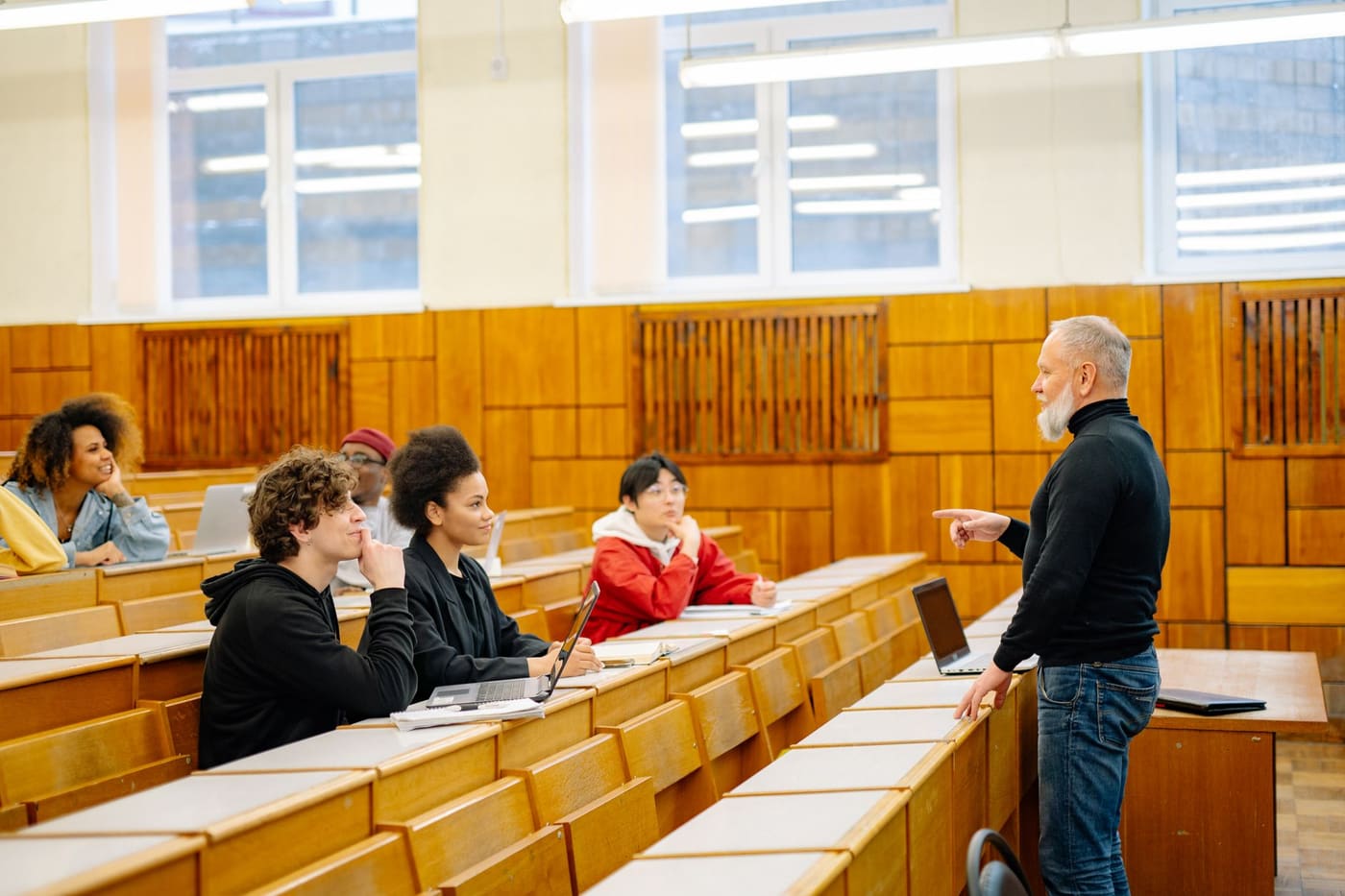 A bearded professor points toward students seated at long wooden desks with laptops in a university lecture hall