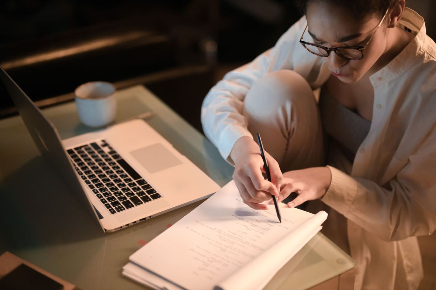 A woman in glasses marks up printed pages beside an open laptop and a coffee cup at night