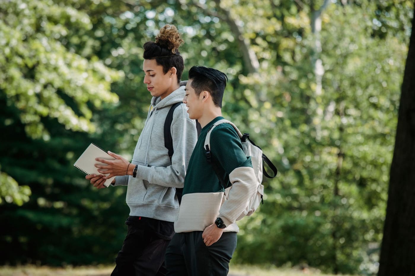 Two graduate students with backpacks walk through a leafy campus while one reads from an open notebook