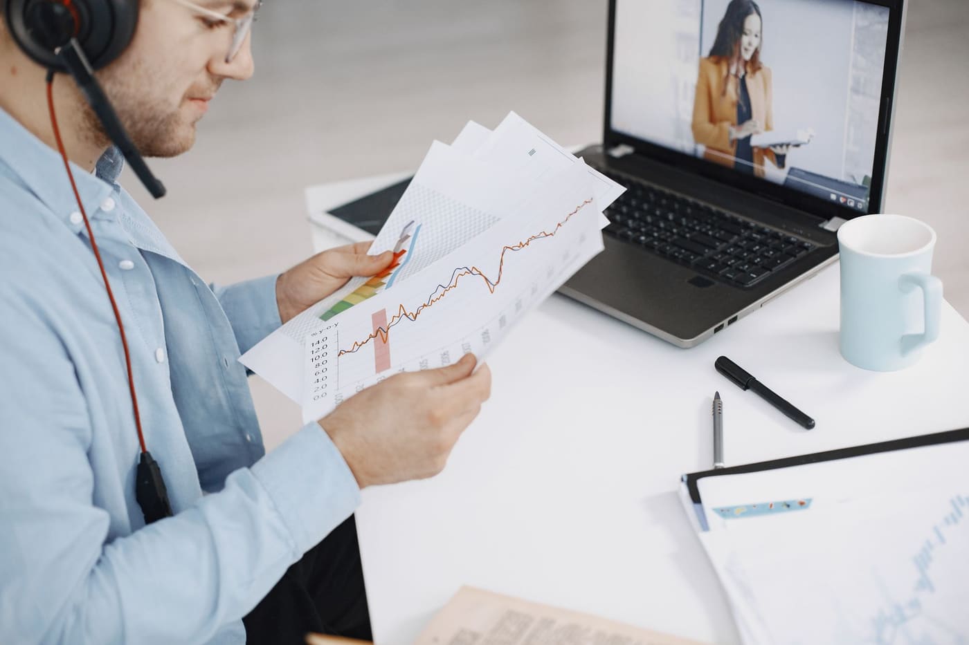 A man wearing a headset reviews printed line charts at a desk while a colleague appears on a video call on his laptop