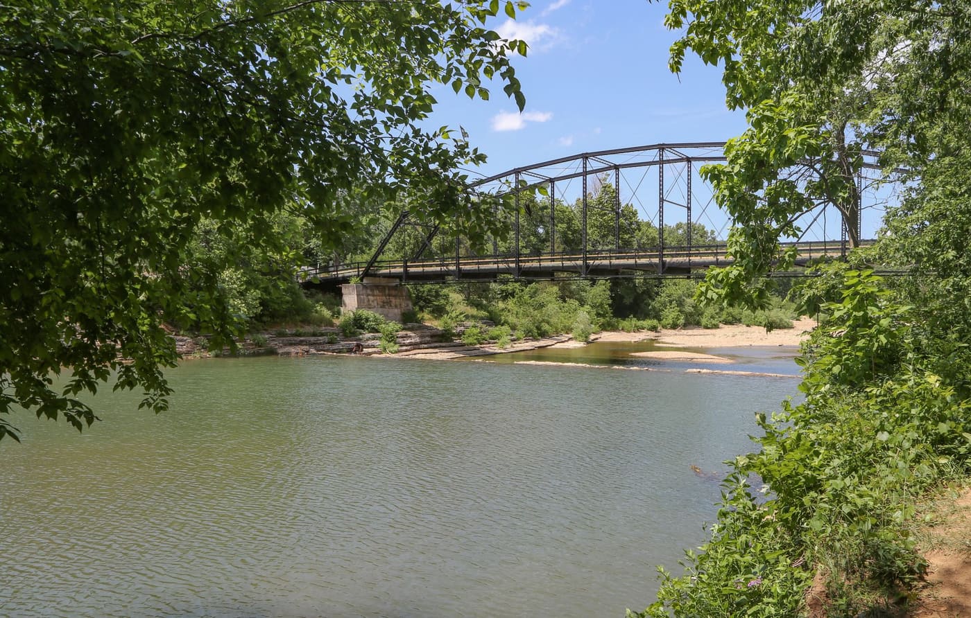 A historic steel truss bridge over a shallow, tree-lined river in the Arkansas Ozarks on a summer day