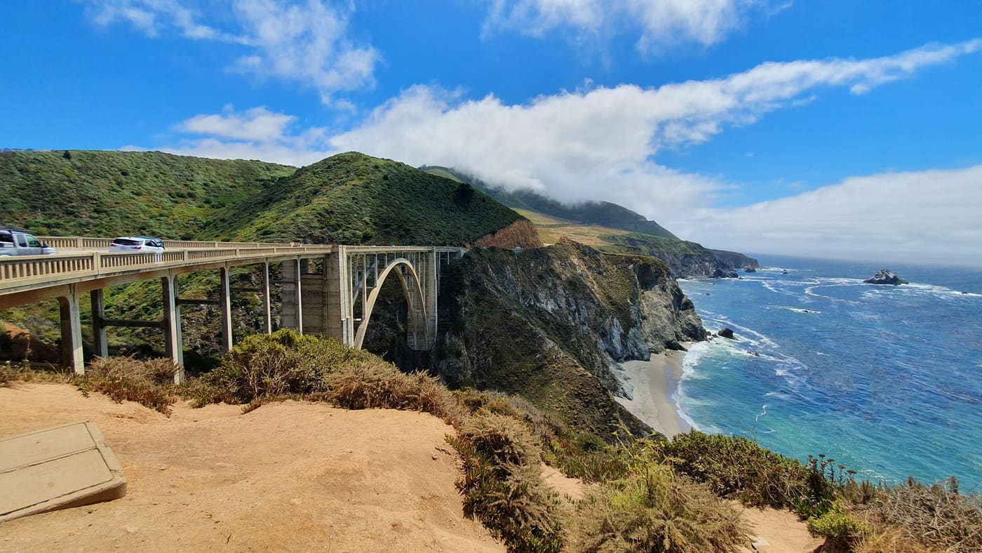 Bixby Creek Bridge arching over a rocky canyon on the Big Sur coast of California, with green hills and blue Pacific surf