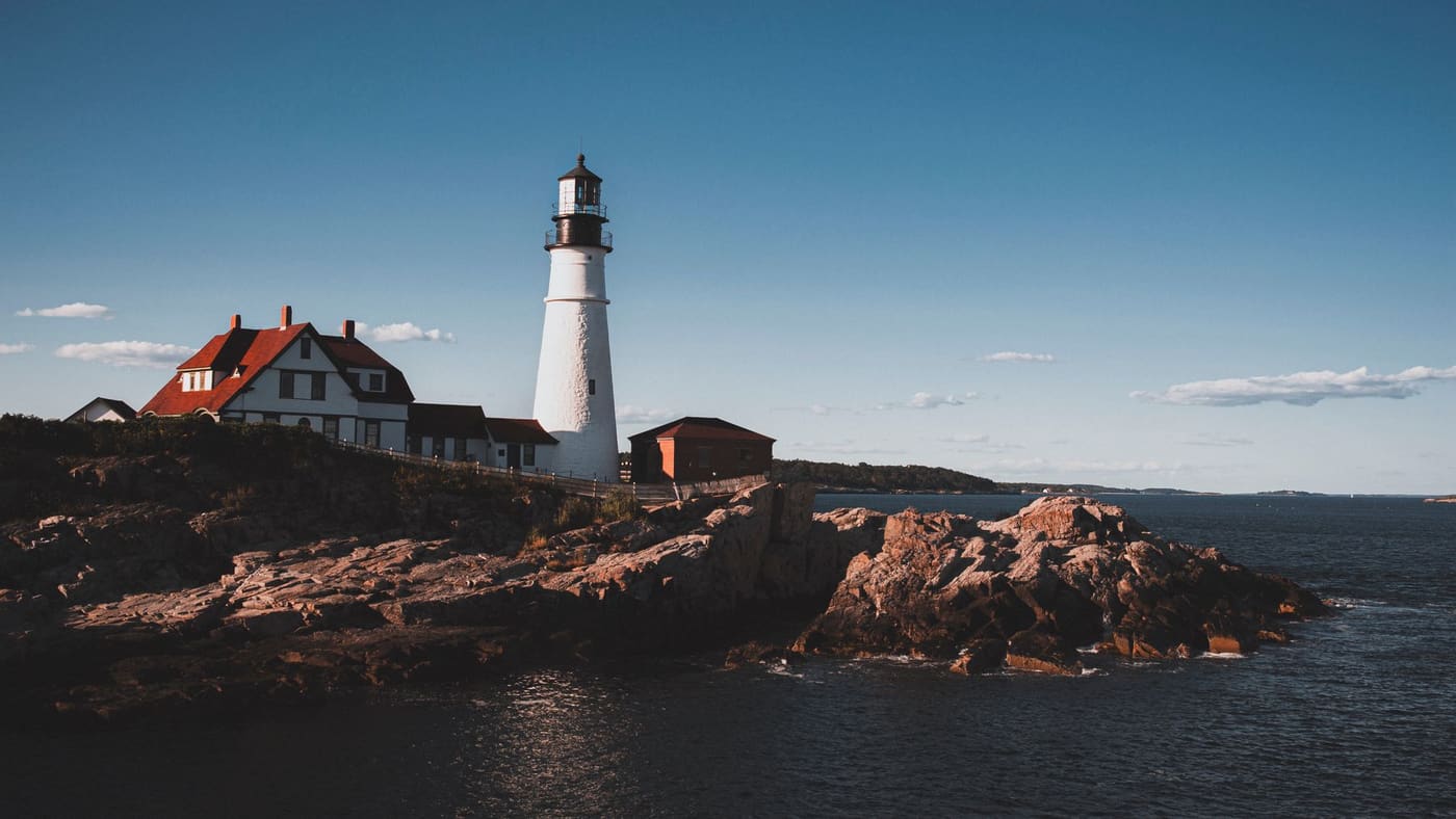 A white lighthouse and red-roofed keeper's house stand on a rocky point above dark water on the Maine coast