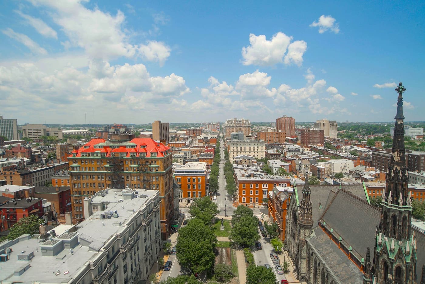 Aerial view down a tree-lined Baltimore avenue with brick rowhouses, an orange-roofed apartment building and a Gothic church spire