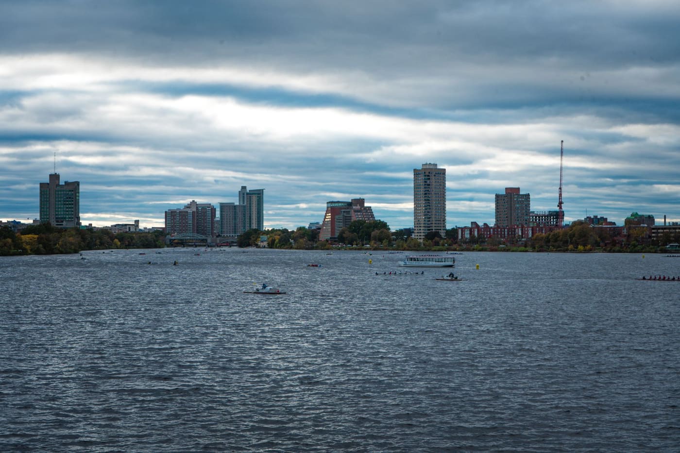 Rowing crews and a tour boat on the Charles River under heavy clouds, with Boston apartment towers along the far bank