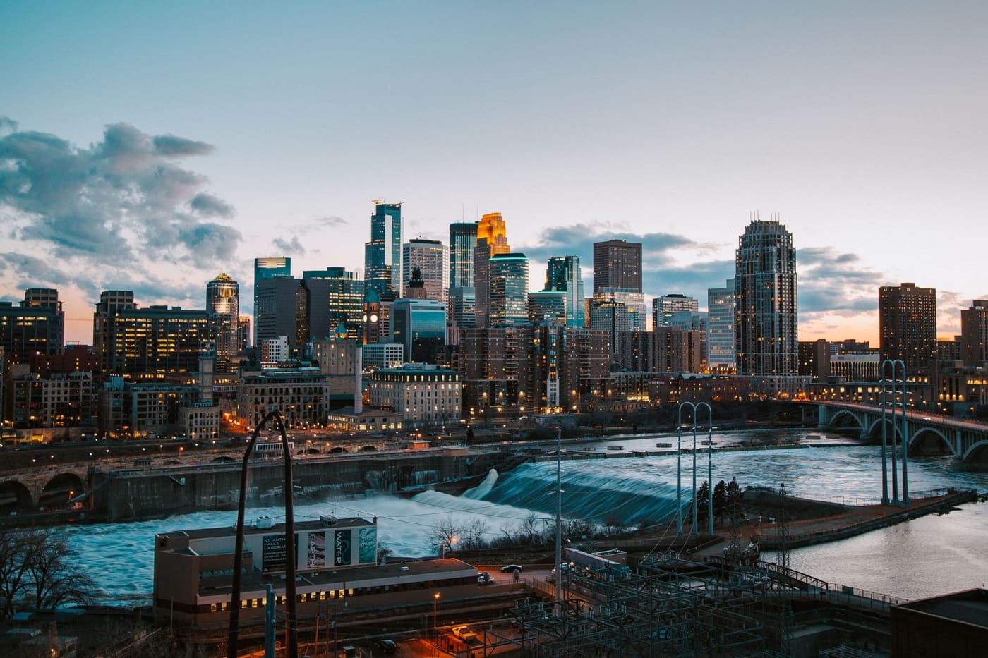 Downtown Minneapolis towers at dusk above St. Anthony Falls and the Mississippi River, with an arched bridge on the right