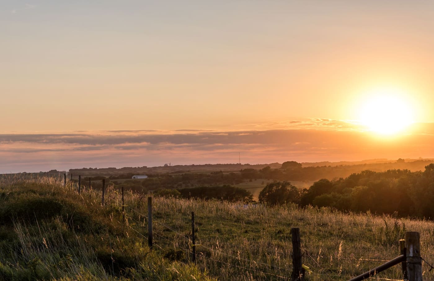 Sun setting over rolling Nebraska pasture, with a barbed-wire fence, tall grass and a distant farm building