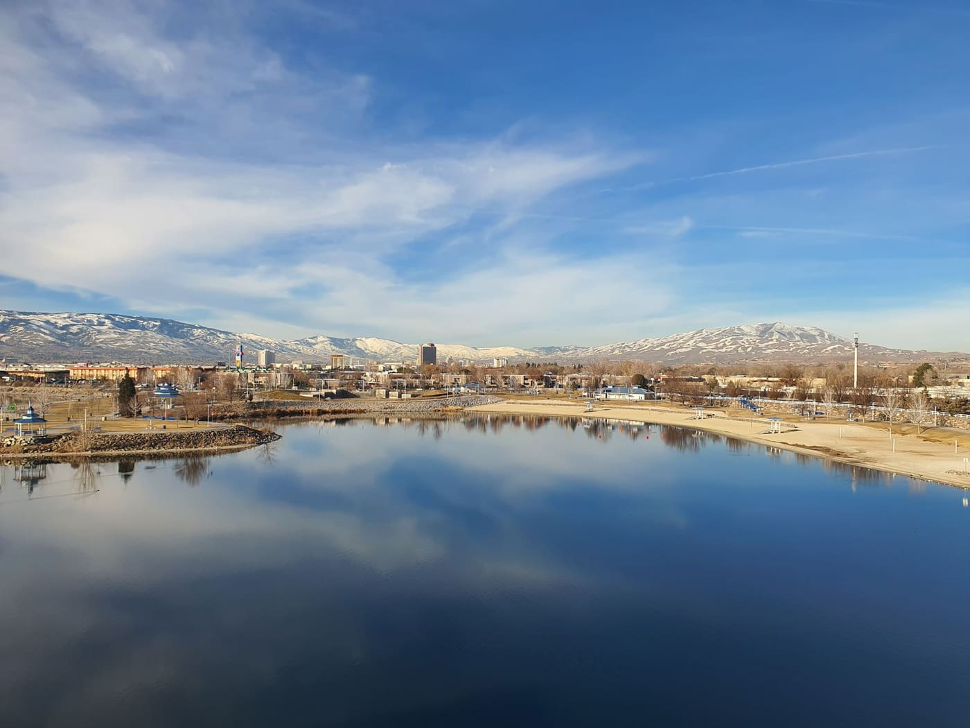 A still lake in a Reno park reflecting the sky, with the downtown skyline and snow-dusted hills behind it