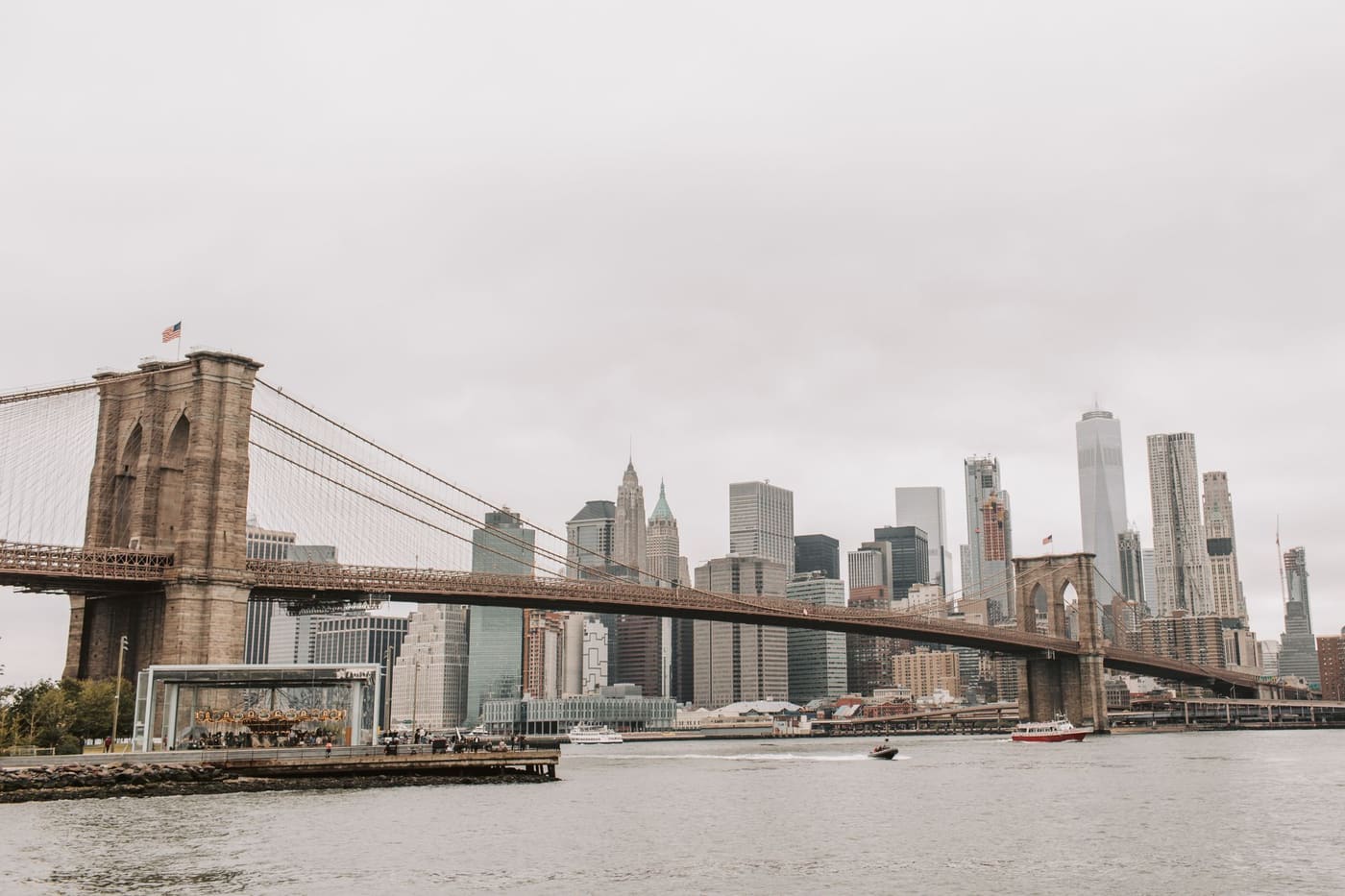 The Brooklyn Bridge spanning the East River toward the Lower Manhattan skyline on an overcast day, with a carousel pavilion on the Brooklyn shore