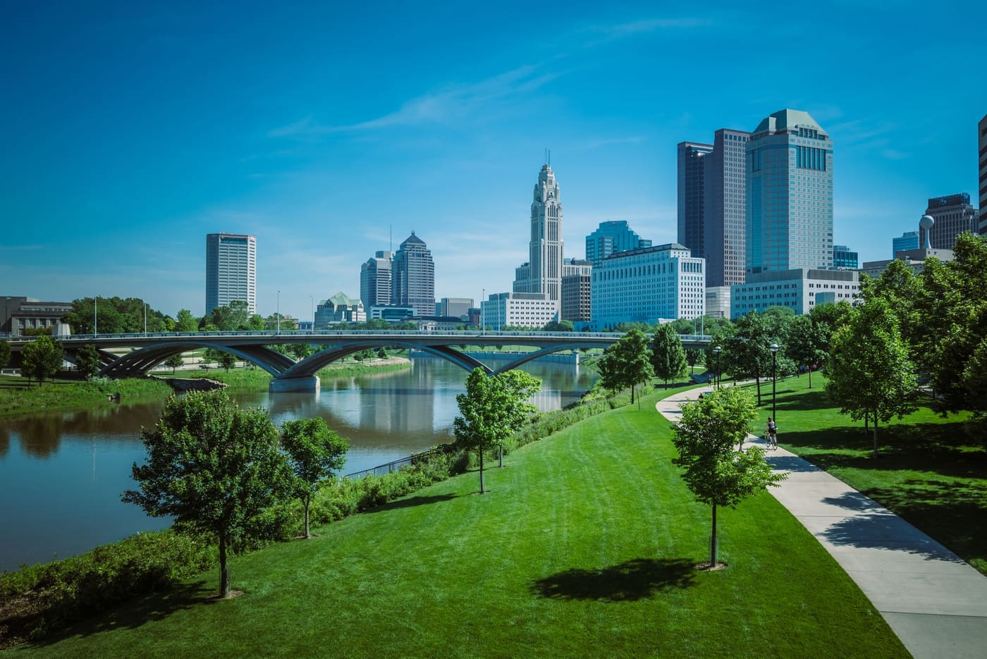 Downtown Columbus skyline and the LeVeque Tower rising behind a bridge over the Scioto River, with a riverside park path in the foreground