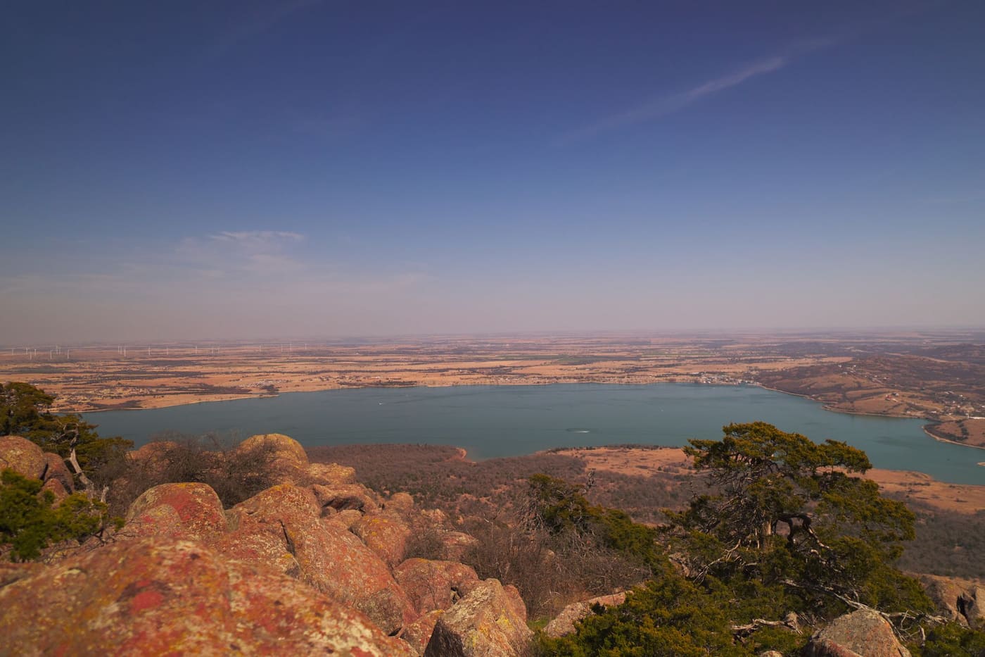 Red granite boulders and cedar trees above Lake Lawtonka in the Wichita Mountains, with flat Oklahoma farmland and wind turbines on the horizon