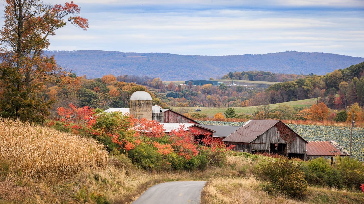 Red barns and a stone silo beside a cornfield and autumn trees, with forested ridges behind, near Williamsport, Pennsylvania