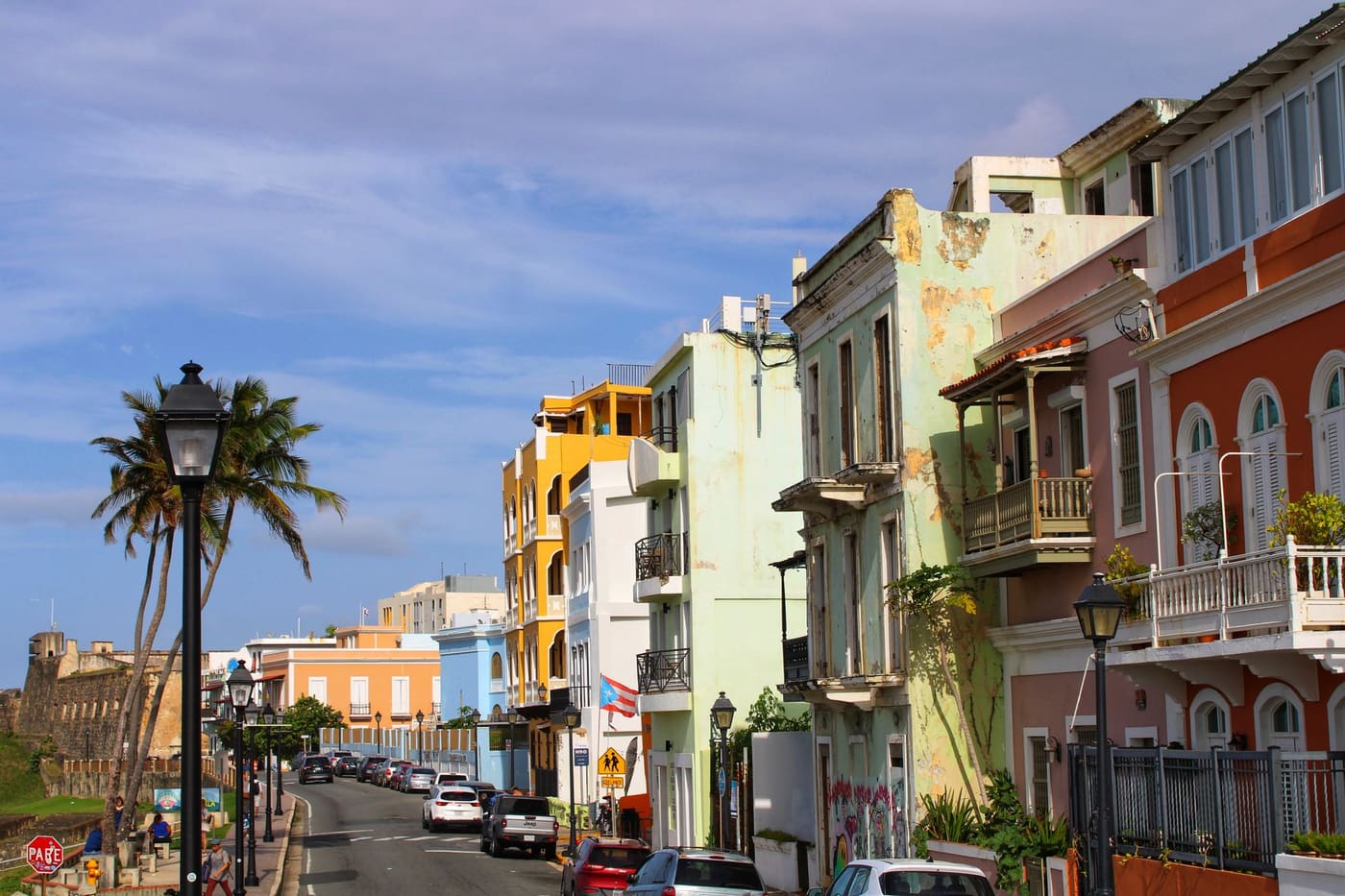 Pastel colonial buildings with balconies line a sunny street in Old San Juan, with palm trees and the old city wall on the left