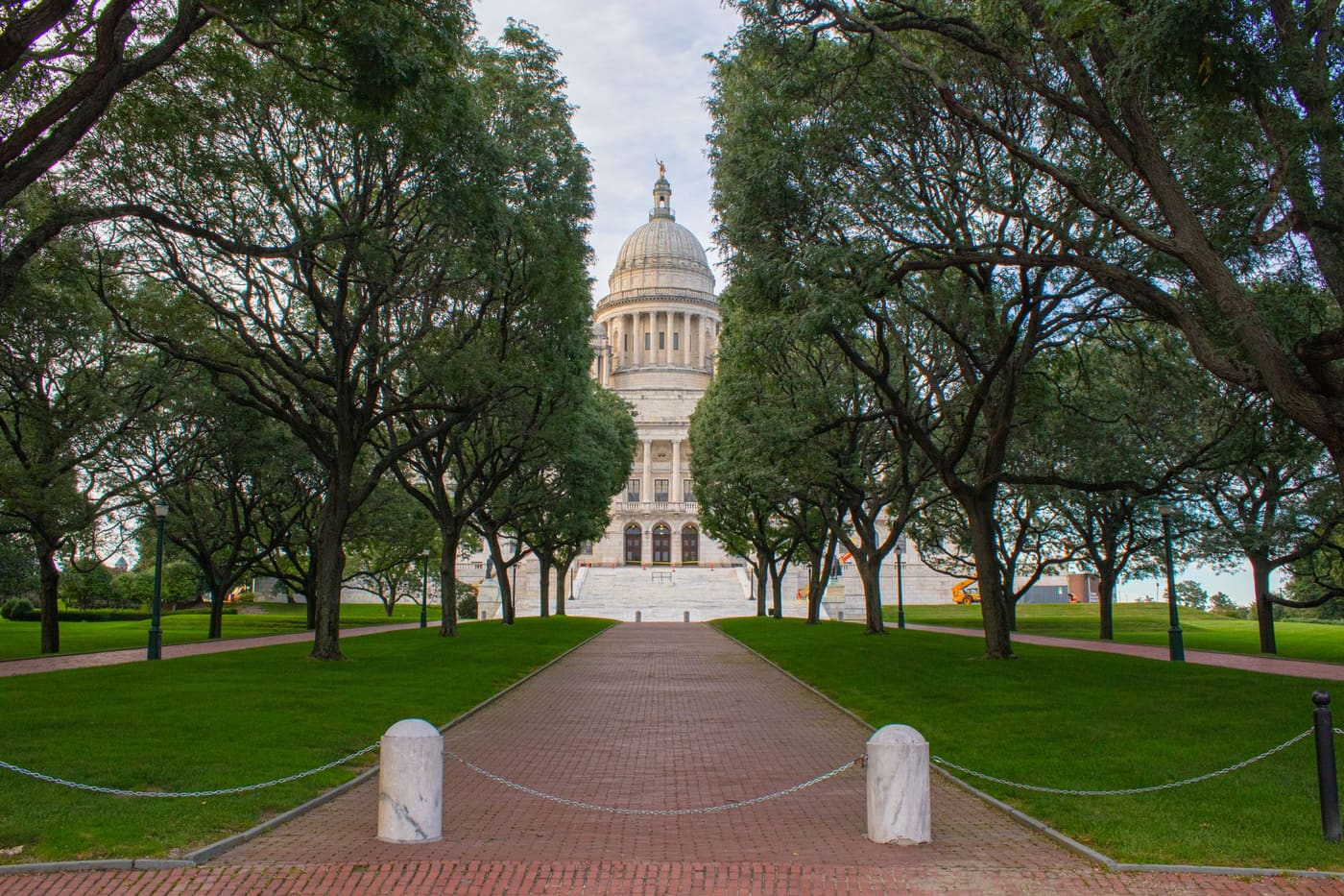 A brick walkway lined with trees leads to the white marble dome of the Rhode Island State House in Providence