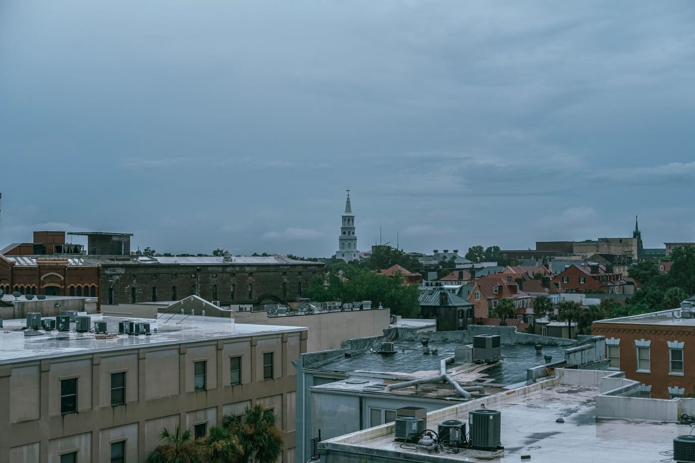 Rooftops of downtown Charleston, South Carolina under a gray sky, with the white steeple of St. Michael's Church rising above brick buildings and palm trees