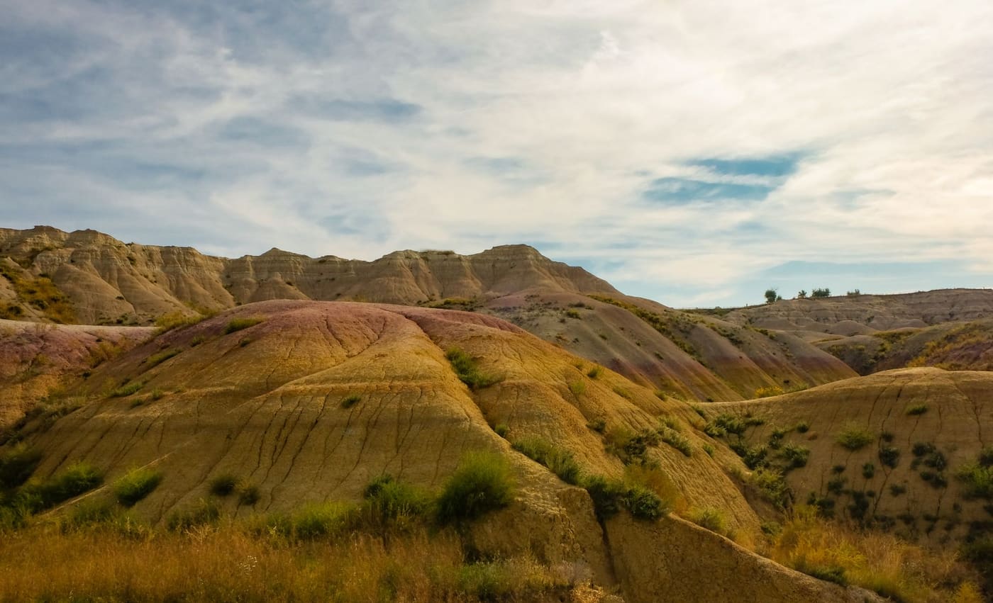 Eroded yellow and rust-colored hills of Badlands National Park in South Dakota under a streaked blue sky, with green shrubs on the slopes