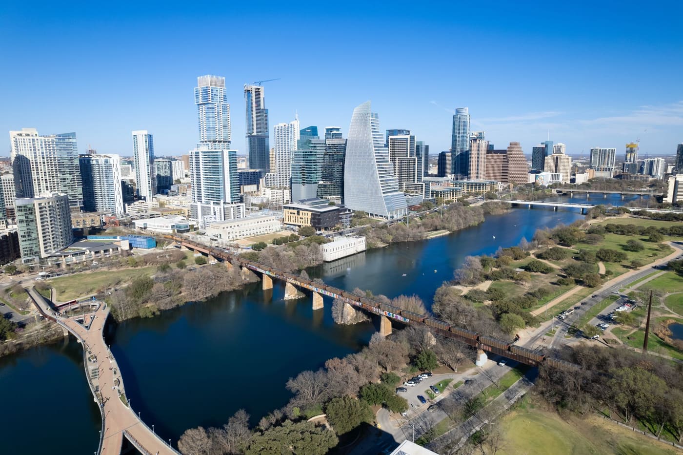Aerial view of downtown Austin's towers above Lady Bird Lake, with a pedestrian bridge and a rail bridge crossing the water