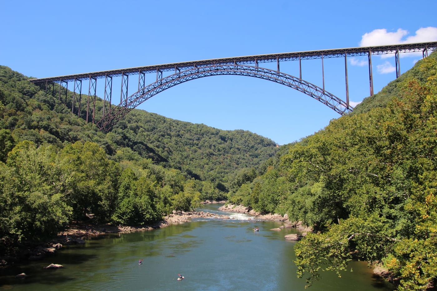 The steel arch of the New River Gorge Bridge spans a forested gorge above the New River, with kayakers on the water below