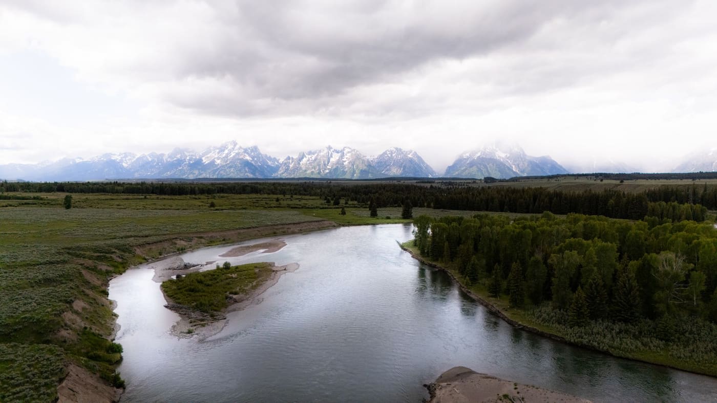 The Snake River bends through green meadows and spruce forest below the snow-streaked Teton Range under low clouds