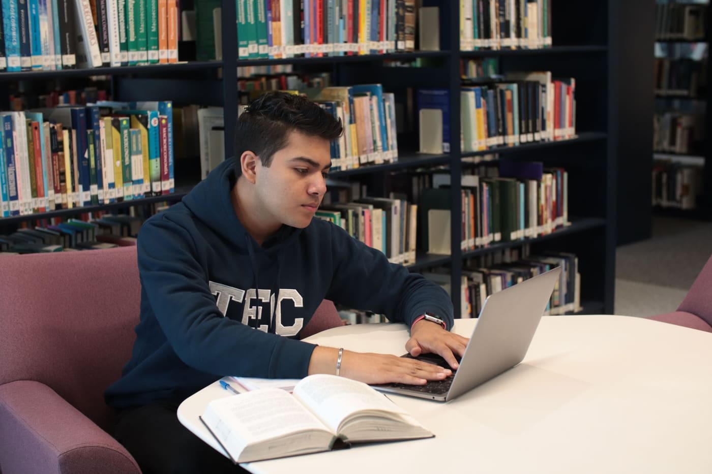 A graduate student working on a laptop at a library table with an open textbook